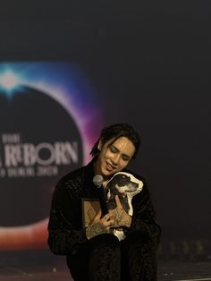 a man holding a small dog in his lap while sitting on a stage with a microphone