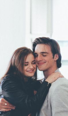 a man and woman hugging each other in front of a white wall with windows behind them