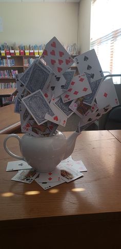 playing cards are falling out of a teacup on a table in front of bookshelves