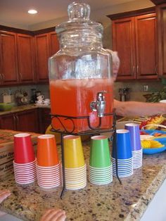 a large jar filled with liquid sitting on top of a counter next to colorful cups