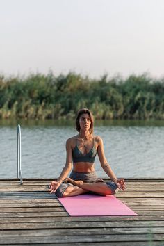 a woman sitting on a pink yoga mat in front of the water with her legs crossed