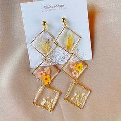 three square earrings with flowers in them on top of a white card and beige fabric