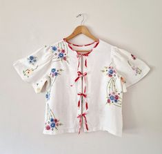 a white blouse with red and blue flowers is hanging on a hanger against a wall