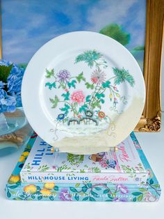 a white plate sitting on top of a stack of books next to a vase with blue flowers