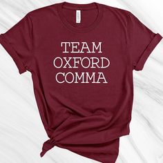 Team Oxford Comma IMPRINT COLOR MAY VARY, DEPENDING ON THE SHIRT COLOR SELECTED. Our solid color unisex tees are super soft ring-spun cotton...it's sure to be your new favorite tee (heather tees are a soft cotton-poly blend)! We use name brands like Bella+Canvas and District. We custom design, print, hand press, and carefully ship everything you see in our shop from our retail location in the Pacific Northwest. All of our items are decorated on quality apparel. This unisex essential tee fits lik School Spirit Cotton Shirt With Slogan, College Cotton Slogan Shirt, Cotton College Slogan Shirt, College Tops With Funny Text And School Spirit, College Tops With Funny Text For School Spirit, College Cotton T-shirt With Funny Text, Funny Text Cotton T-shirt For College, Cotton College Shirt With Name Print, Cotton College Shirt With Letter Print