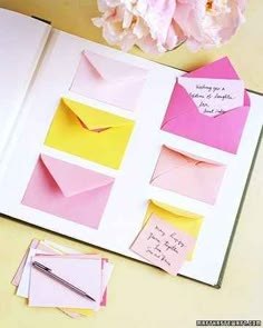 an open book with pink and yellow envelopes on it, next to some flowers