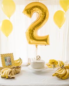 a white table topped with bananas and a number two cake next to balloons in the air