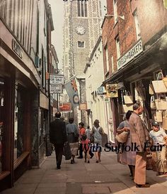 an old photo of people walking down the street in front of shops and buildings with a clock tower behind them