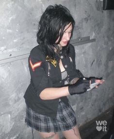 a woman in a black shirt and plaid skirt holding a cell phone while standing next to a wall