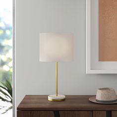 a lamp on a wooden table next to a white wall and framed photograph in the corner