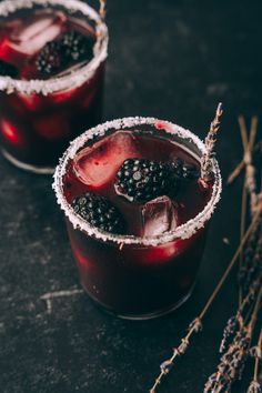 two glasses filled with blackberries and ice on top of a dark table next to dried plants