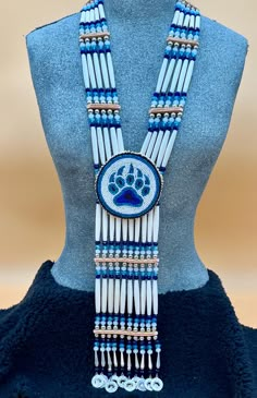 Bone hairpipes and crystal beads are often seen as part of traditional Native American dancers.  This modified breastplate  has been created to proudly proclaim that this is a good day to be Indigenous . It is designed to be worn not only in the dance circle, but in mainstream life, showing your Native pride and fashion sense.   The necklace is 10 vertical strands of bone hairpipes, nickel beads and 8mm sparking, faceted, fire-polished glass crystal beads in the colors of navy blue, turquoise blue and white frost The centerpiece is a beaded rosette of a Bear paw in the same blue colors on a white background. At the bottom of breastplate, pewter and rhinestone pendants  add their music to your movements. The breastplate comes with matching earrings on hypoallergenic lever back wires featuri Medallion Necklace Beaded, Native American Medallions, Powwow Regalia Jingle Dress, Native Medallion, Powwow Beadwork, Indigenous Fashion, Native American Dance, American Dance, Creative Fabric