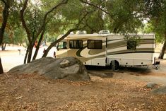 an rv parked in the woods next to a large rock and trees with people standing around it