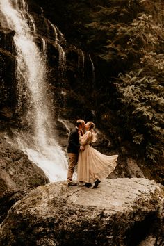 a bride and groom standing on a rock in front of a waterfall with their arms around each other