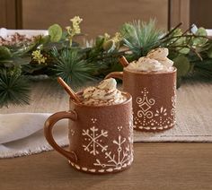 two mugs with hot chocolate and whipped cream in them sitting on a table next to evergreen branches