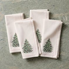 three embroidered christmas trees on white linen napkins sitting on a stone counter top with red stitching