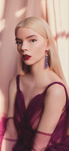 a woman with long blonde hair wearing purple dress and large earrings, leaning against a wall