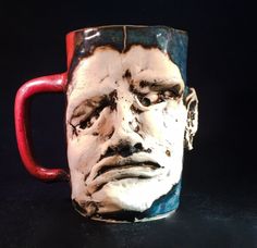 a ceramic mug with a face painted on it's side and red handle, sitting on a black surface