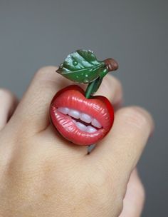 a woman's hand holding a ring with a cherry on it and a green leaf sticking out of the middle