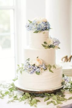 a three tiered white cake with blue flowers on the side and greenery around it