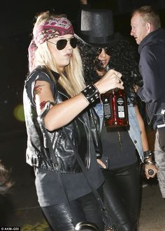 a woman in leather pants and a top hat is holding a bottle while another man looks on