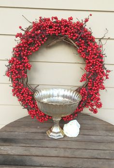 a metal bowl with red berries on it sitting on a wooden table next to a wreath