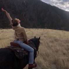 a woman riding on the back of a brown horse in a field next to a mountain