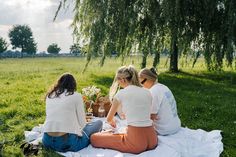 three women are sitting on a blanket in the grass and having a picnic under a willow tree