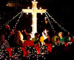 a group of people sitting on top of a cross covered in christmas lights at night