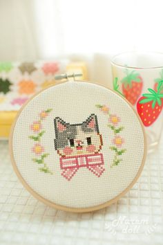 a cross - stitch cat is sitting on a table next to a cup and strawberries