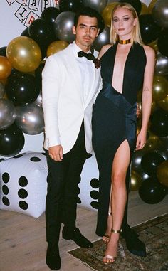 a man and woman standing next to each other in front of balloon wall with black and gold balloons