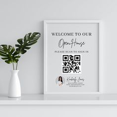 a white vase with a green plant in it next to a framed poster that says welcome to our open house please scan to sign in