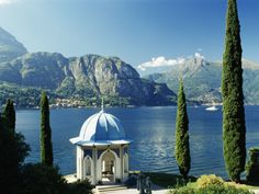 a gazebo in the middle of a lake surrounded by trees and bushes with mountains in the background