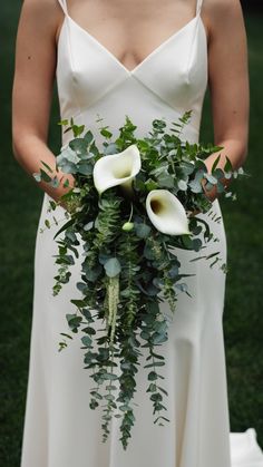 a woman in a white dress holding a bouquet of greenery and calla lilies