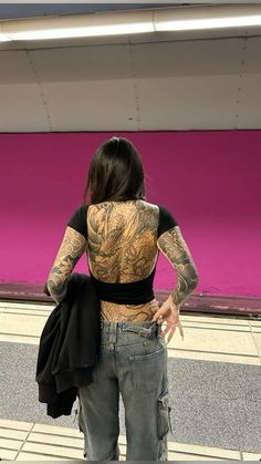 a woman standing in front of a pink wall with tattoos on her back and arms