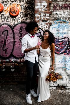 a man and woman standing next to each other in front of a wall with graffiti