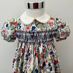 This one of a kind dress features hand-smocking and embroidery, on beautiful quality floral cotton lawn fabric with contrasting  collar featuring picot edge.   This fabric is beautiful quality, fine and light-weight so cool and comfortable for little ones to wear. The full-skirted dress has a generous hem and back sashes tied in a bow, to maximise the seasons of wear. Fits up to 54cm chest measurement.   Length is approximately 50cm from shoulder to hem, although there's plenty of hem allowance Multicolor Smocked Dress With Ruffles For Garden Party, Multicolor Smocked Dress For Garden Party, Multicolor Smocked Dress With Smocked Back For Garden Party, Cream Smocked Bodice Dress For Garden Party, Multicolor Cotton Smocked Dress With Floral Print, Multicolor Floral Print Cotton Smocked Dress, Cream Dress With Smocked Bodice For Garden Party, Multicolor Floral Dress With Smocked Bodice For Garden Party, Multicolor Floral Dress For Garden Party