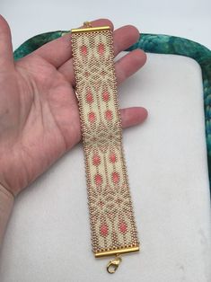 a hand is holding a gold and red beaded bracelet on a white cloth background