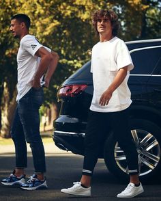 two young men standing next to each other in front of a black car on the street