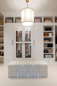 a closet with white cabinets and clothes hanging on the doors, an upholstered bench in front of it