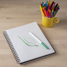 an open notebook with markers and pens next to a cup of pencils on a table