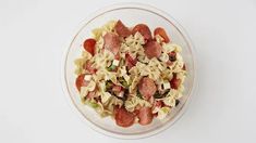 a glass bowl filled with pasta salad and pepperoni on top of a white table