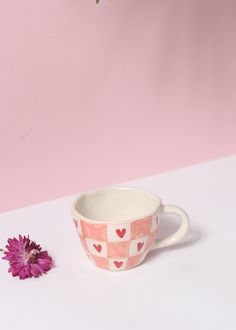 a pink and white cup with hearts on it next to a purple flower in front of a pink wall