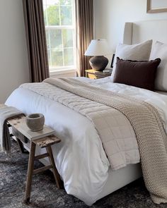 a bed with white sheets and pillows next to a lamp on a table in front of a window