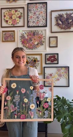 a woman holding up a painting in front of a wall full of pictures and flowers