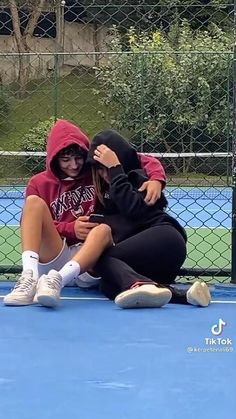 two people sitting on the ground in front of a tennis court with one person covering their face