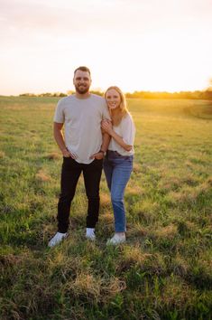 a man and woman standing in the middle of a field at sunset with their arms around each other