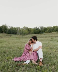 a man and woman sitting on the ground in a grassy field with trees behind them