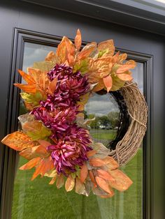 a wreath with purple and orange flowers hanging on a front door window sill in the grass