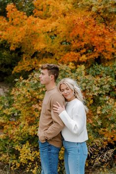 a man and woman standing next to each other in front of trees with orange leaves